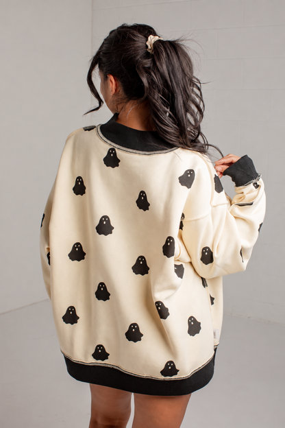 A girl viewed from behind wearing a cream-colored sweatshirt with black ribbed trim and a black ghost pattern, set against a plain light gray wall.