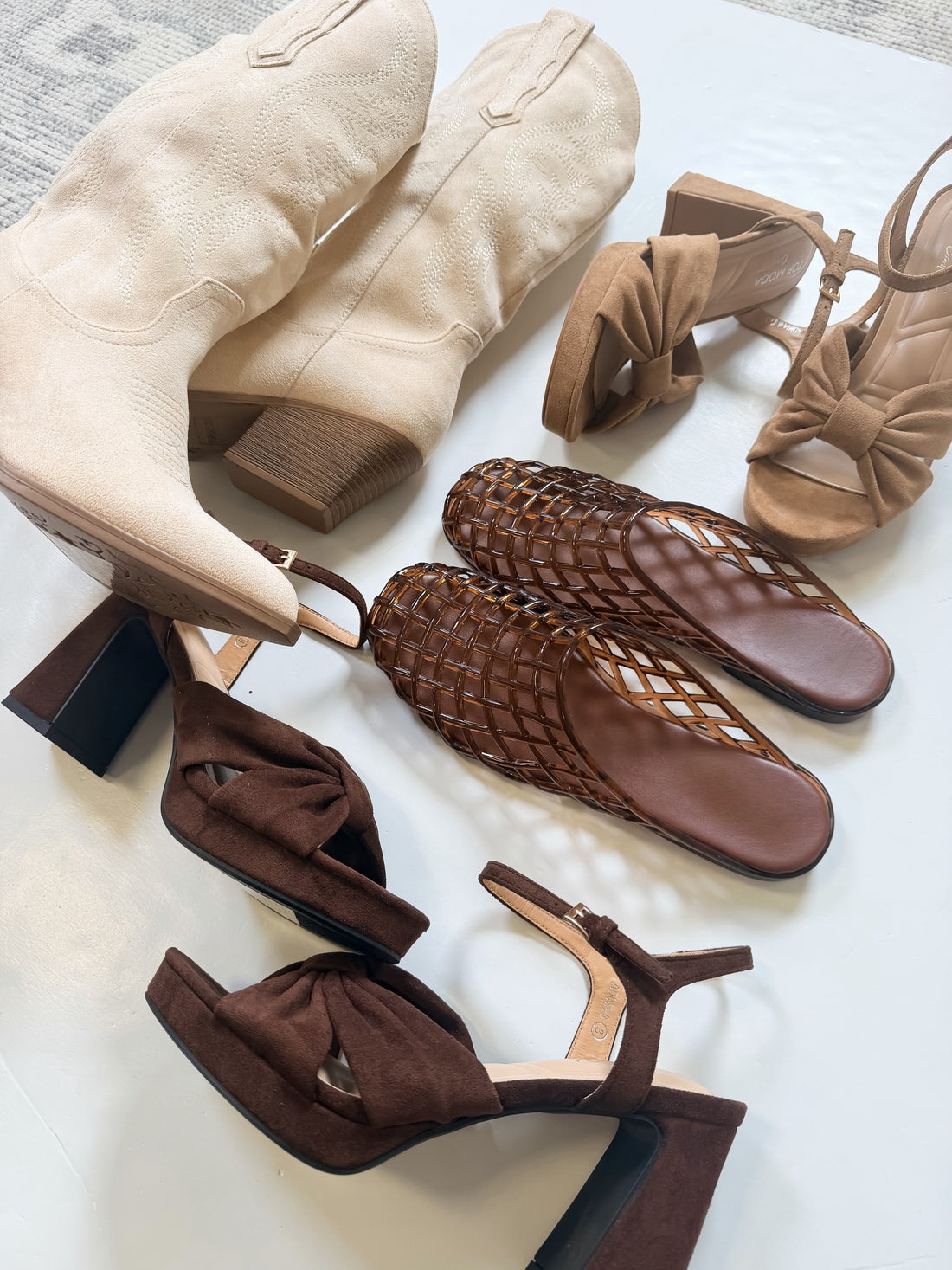 A styled flat-lay of multiple women's heeled shoes in neutral tones, including beige suede ankle boots, tan bow-strap sandals, brown woven mule sandals, and dark brown slingback heels, arranged on a white surface.