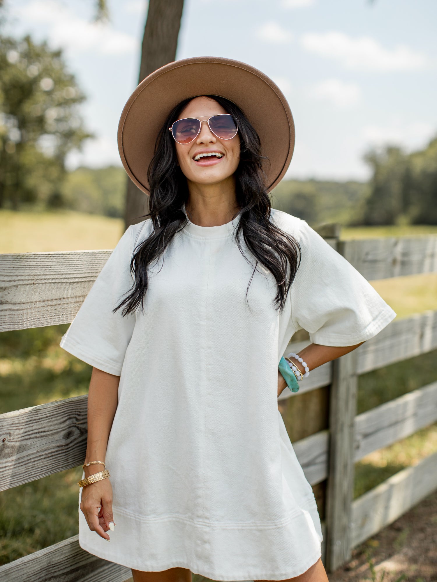 A woman wearing a white short-sleeve Denim tunic dress, a wide-brim tan hat, and sunglasses, standing outdoors in front of a wooden fence.