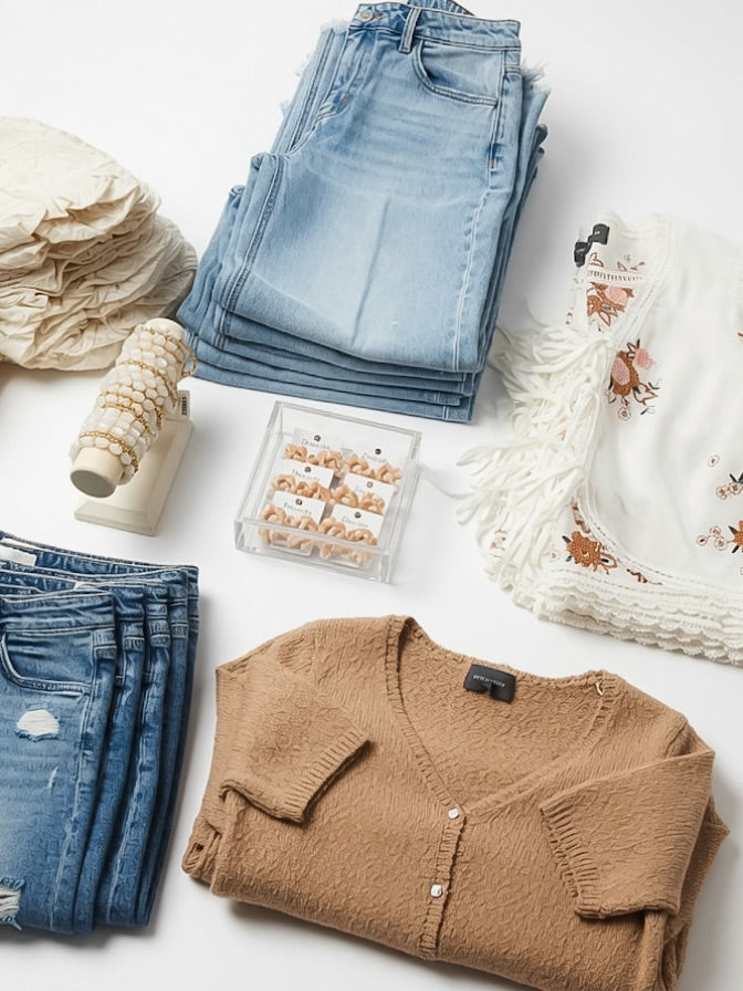 A styled flat-lay of folded clothing and accessories on a white background, including light and dark blue jeans, a tan knit cardigan, a cream fringed top with floral embroidery, a stack of cream ruffled fabric, a small clear jewelry box, and a coiled cream textile.