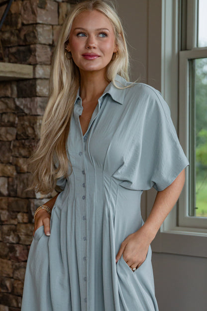 Woman wearing a light blue dress standing in a room with stone walls and a window.