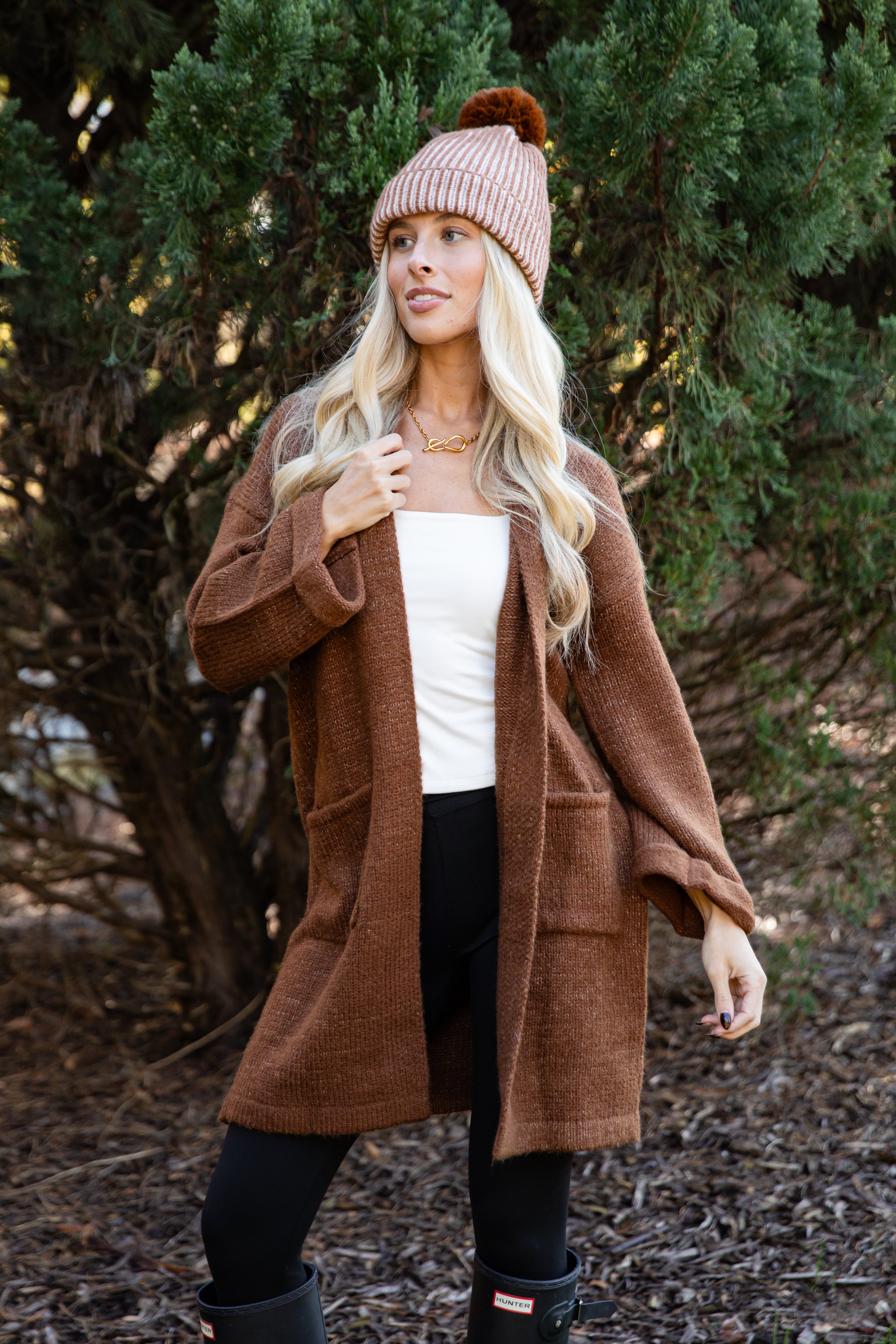 Woman wearing a brown coat, white top, black pants, and a pink beanie outdoors.