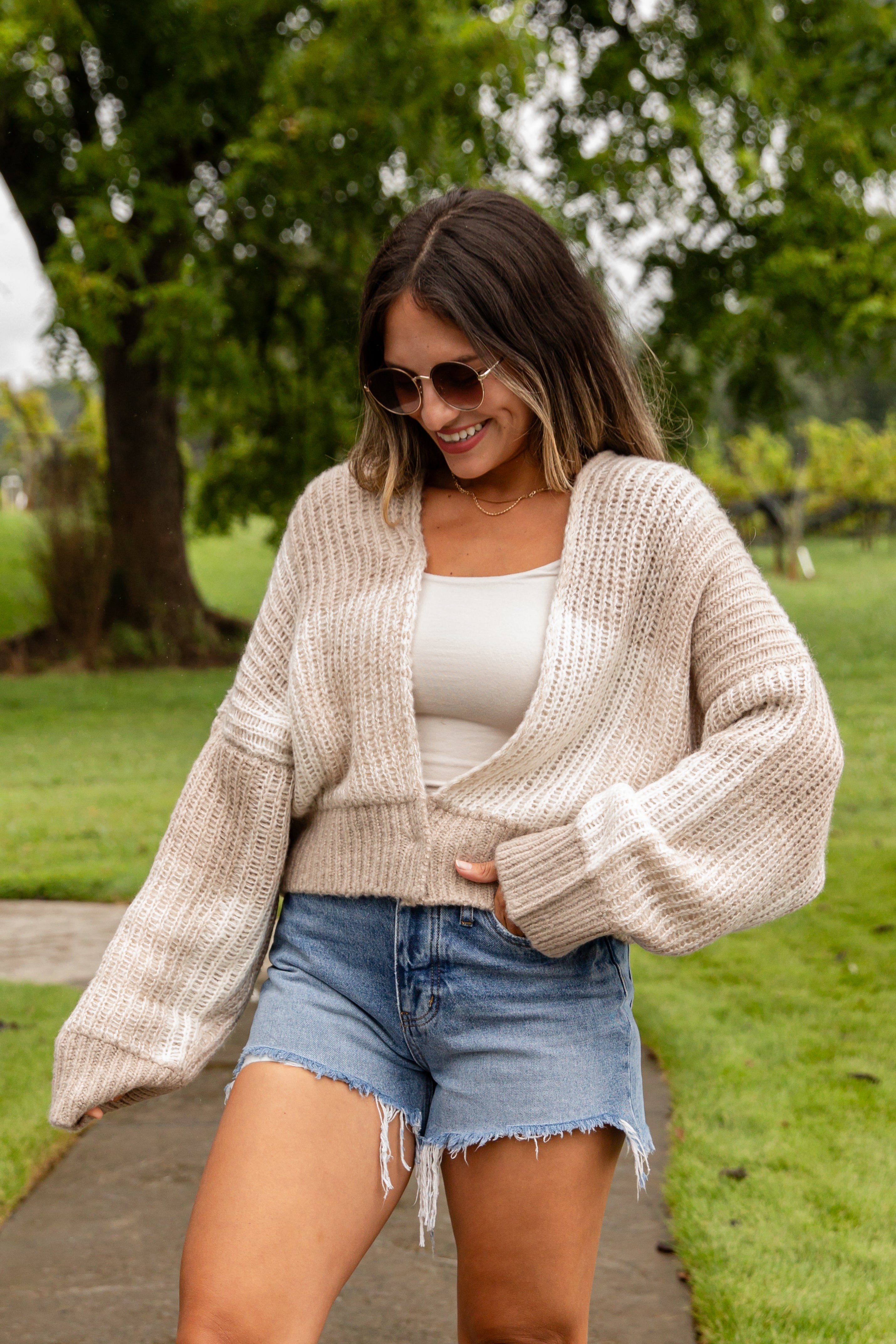 Woman wearing a beige cardigan, white top, and denim shorts outdoors.