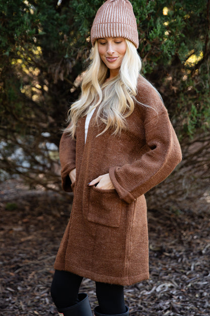 Woman wearing a brown coat and pink beanie standing outdoors with trees in the background