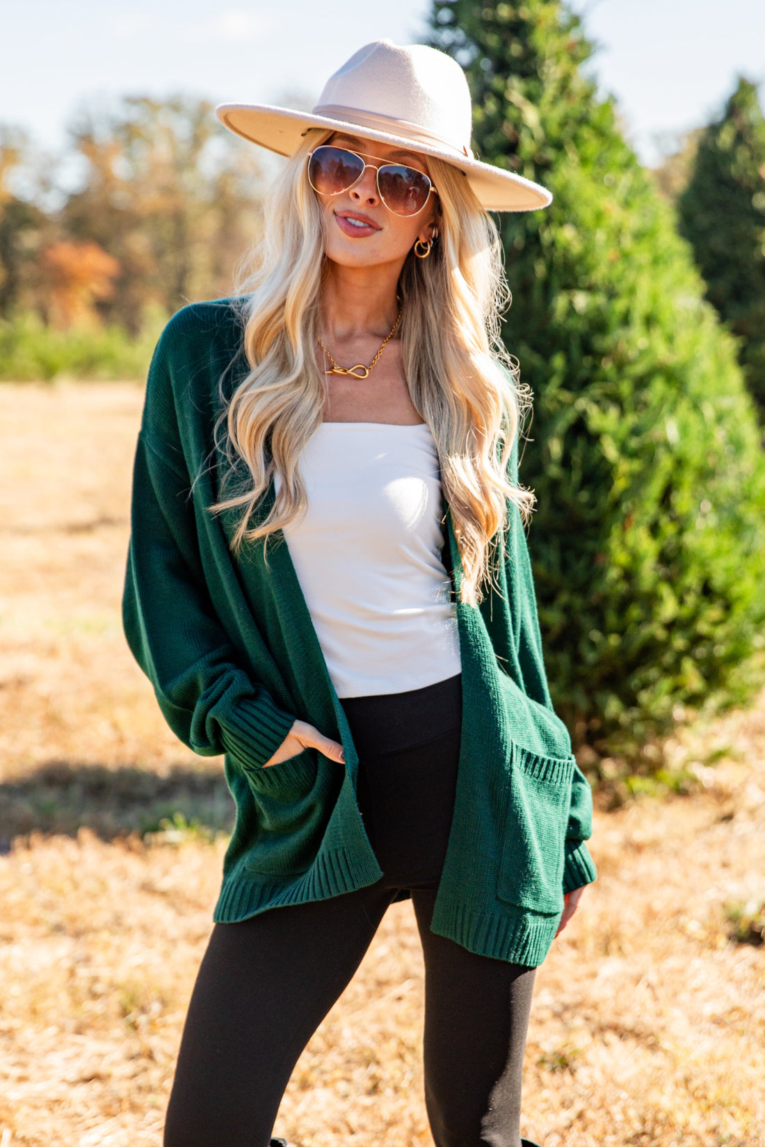 Woman in a green cardigan, white top, black leggings, and wide-brimmed hat standing in a field with trees in the background.