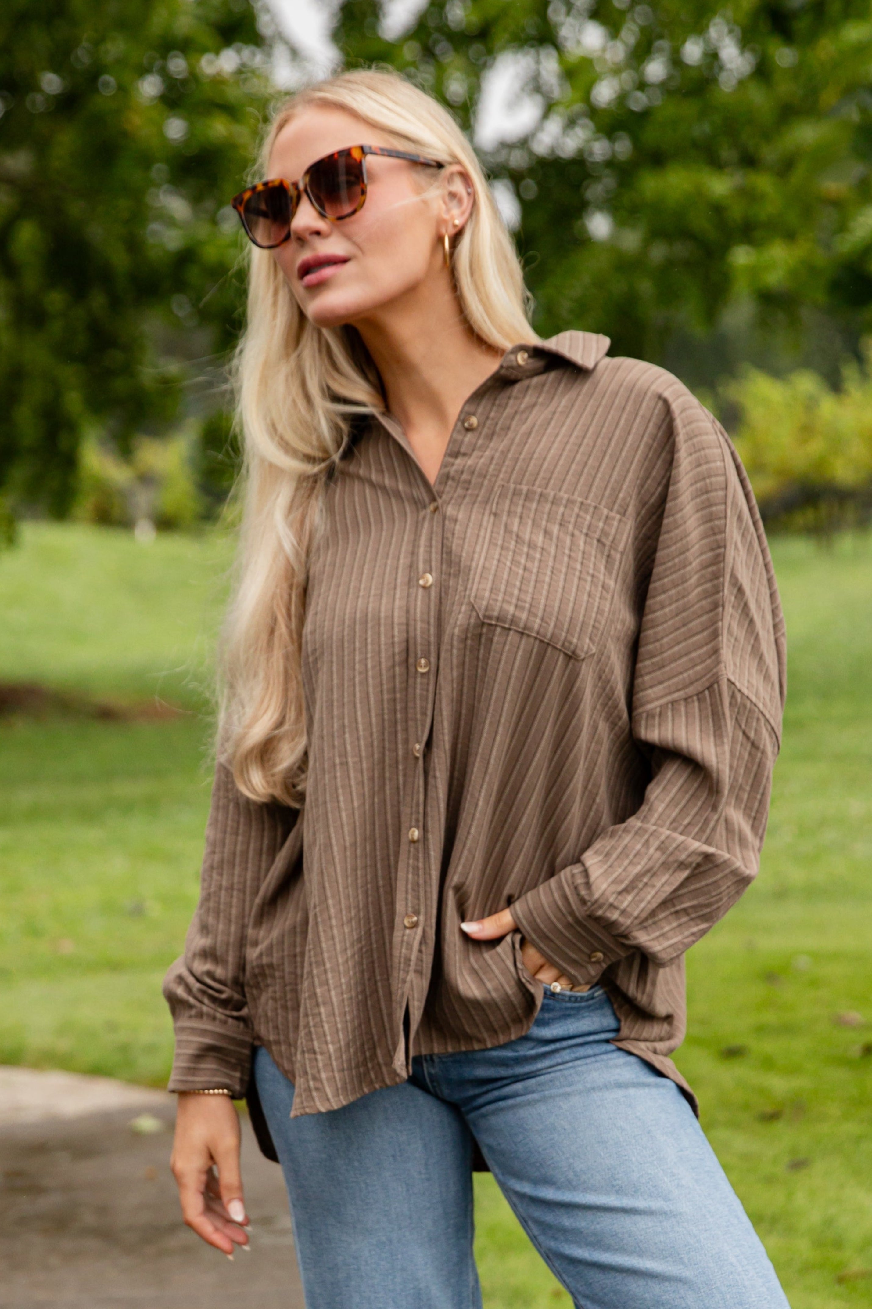 Woman in a brown shirt, blue jeans, and white boots standing on a sidewalk with greenery in the background
