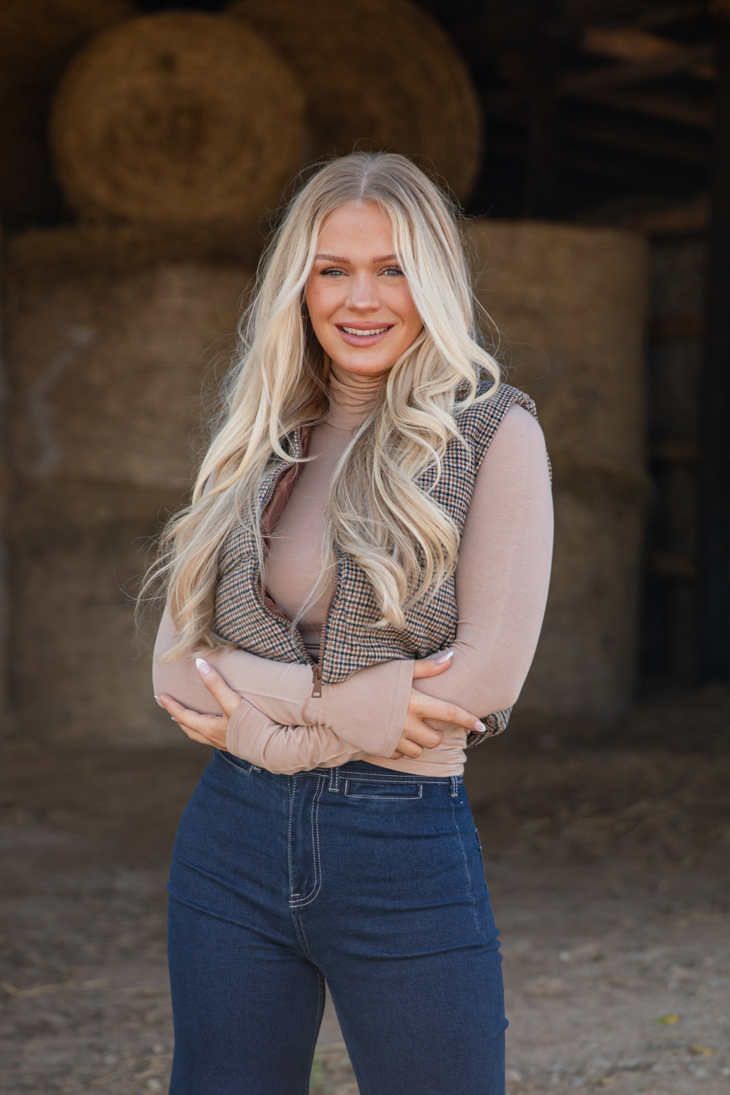 Woman standing in front of hay bales wearing a plaid vest and blue jeans.