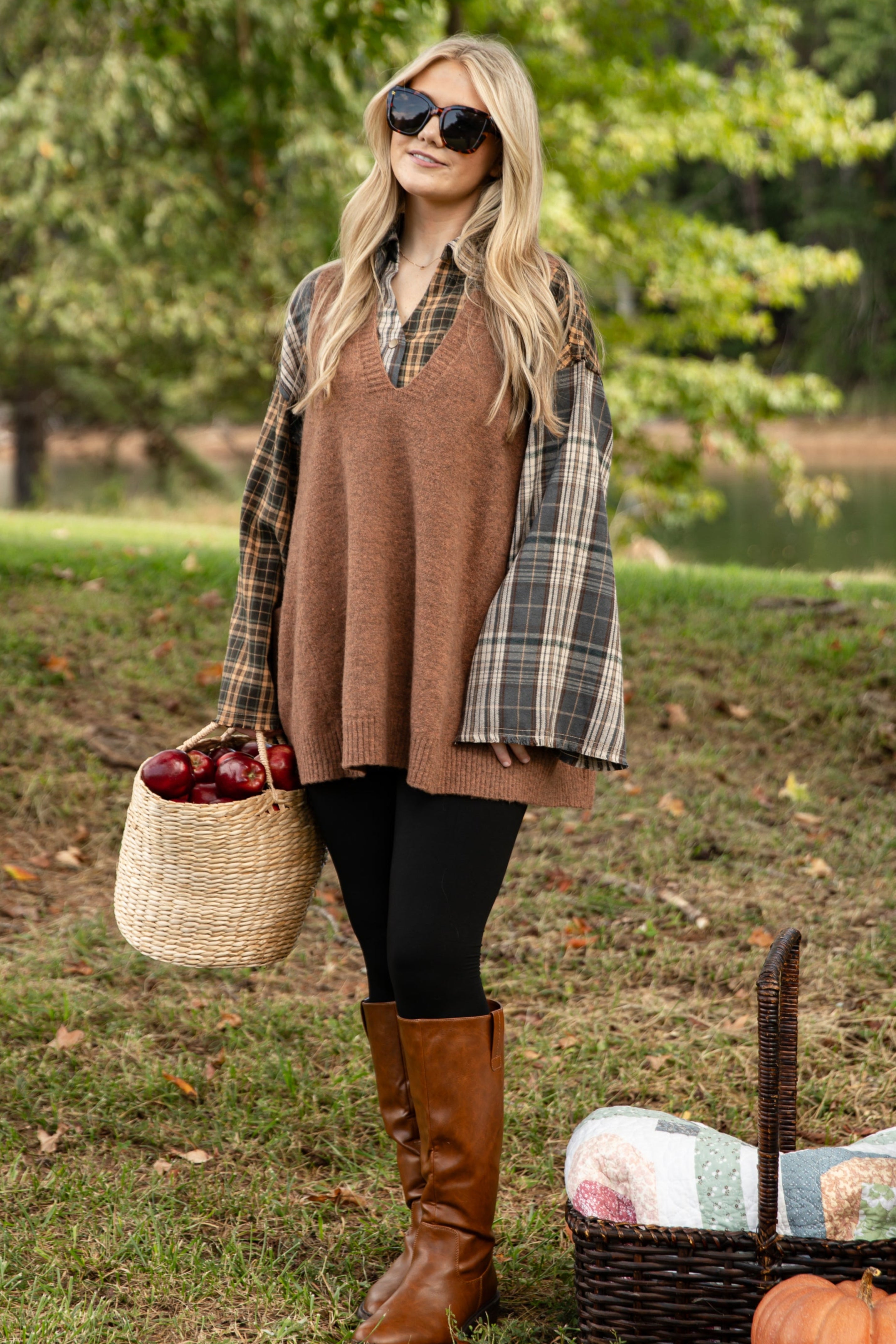 Woman in a plaid shirt and brown sweater standing outdoors with a picnic setup.