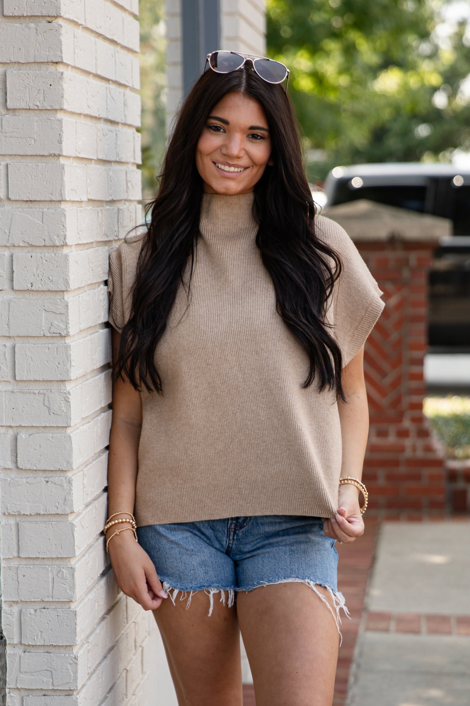 Woman in beige sweater and denim shorts standing against a brick wall.