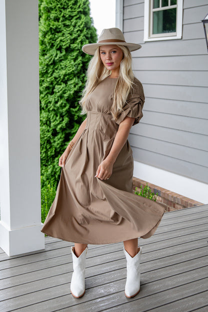 Woman in a beige dress and hat standing on a wooden deck.