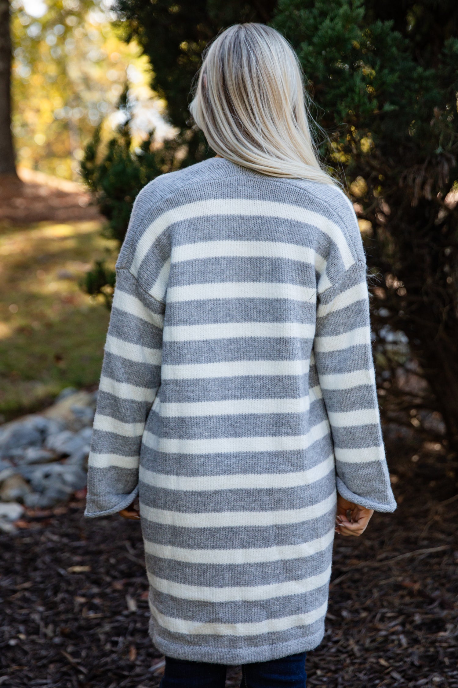 Person wearing a long, striped cardigan in a natural setting with trees and ground cover.