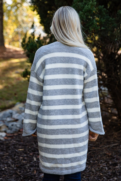 Person wearing a long, striped cardigan in a natural setting with trees and ground cover.
