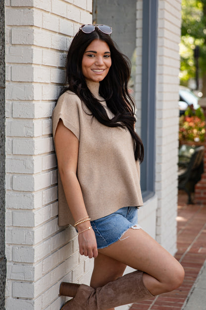 Woman leaning against a brick wall wearing a beige top and denim shorts.