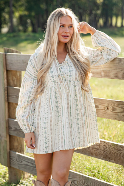 Woman in a white dress and beige boots standing by a wooden fence in a field.