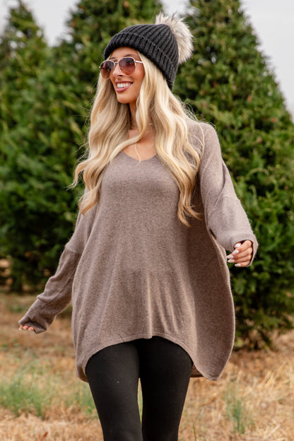 Woman in a brown sweater and black leggings walking through a field with trees in the background