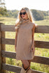 Woman wearing a beige sleeveless sweater dress standing by a wooden fence in a field.