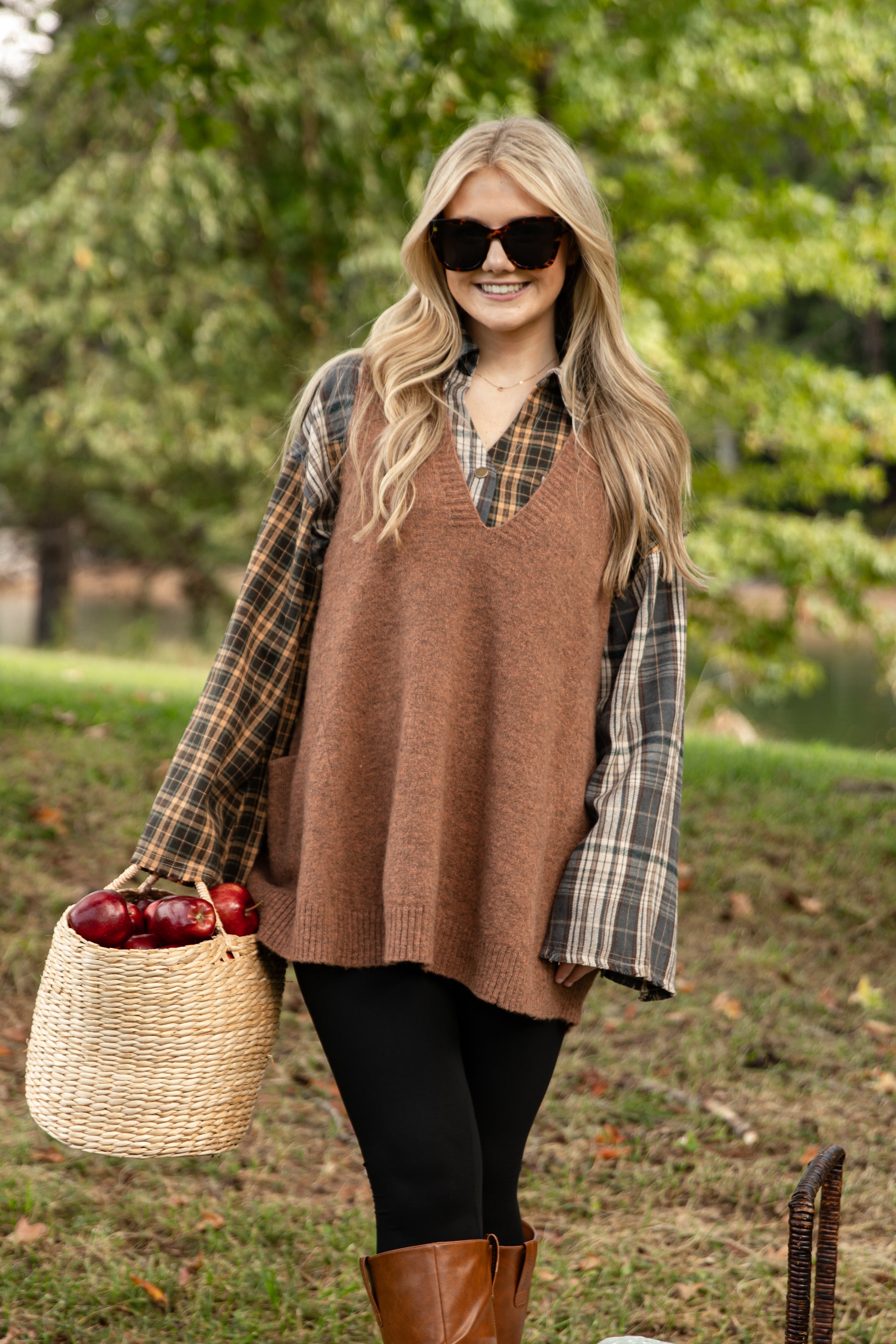 Woman in a brown sweater vest and plaid shirt holding a basket of apples outdoors.