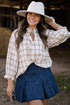 Woman wearing a plaid shirt, denim skirt, and white hat in a rustic setting.