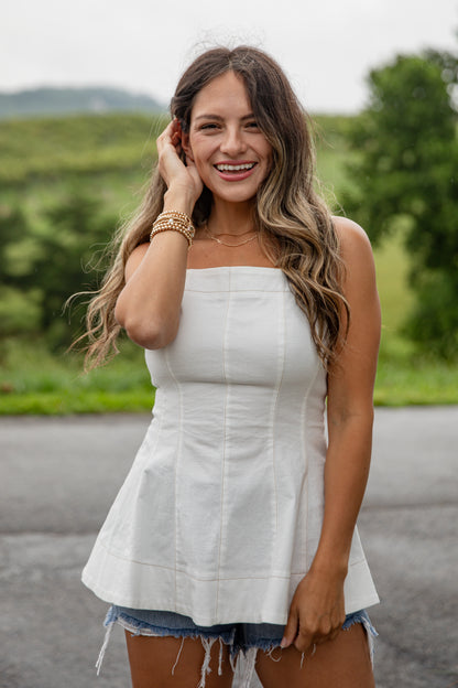 Woman in a white strapless top and denim shorts standing outdoors with greenery in the background