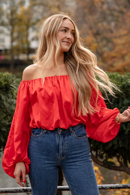 Woman wearing a red off-shoulder top and blue jeans standing in an autumn park.