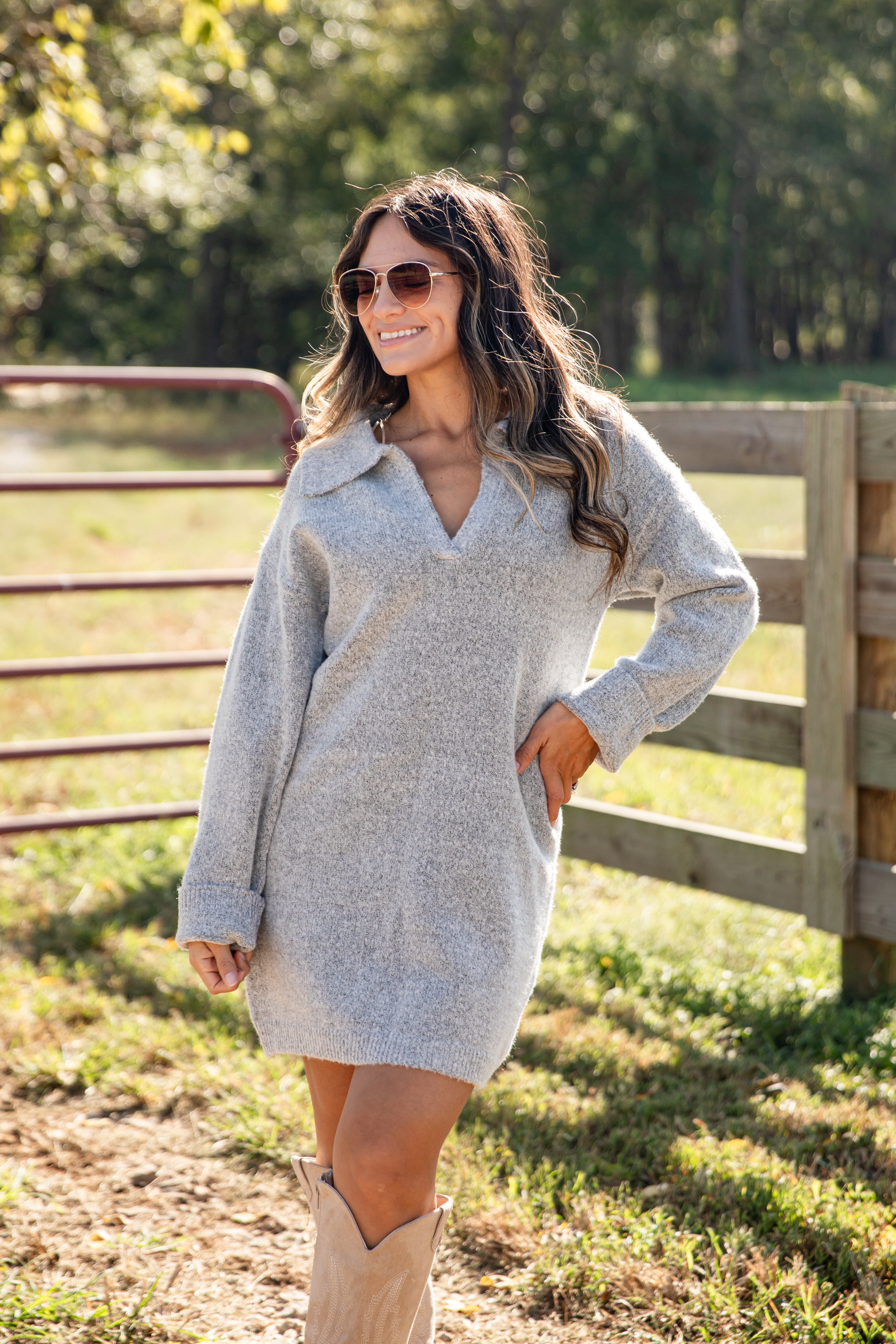 Woman wearing a gray sweater dress and beige boots standing in a field with a wooden fence and trees in the background.