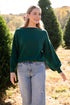 Woman in a green sweater and jeans standing in a field with trees in the background
