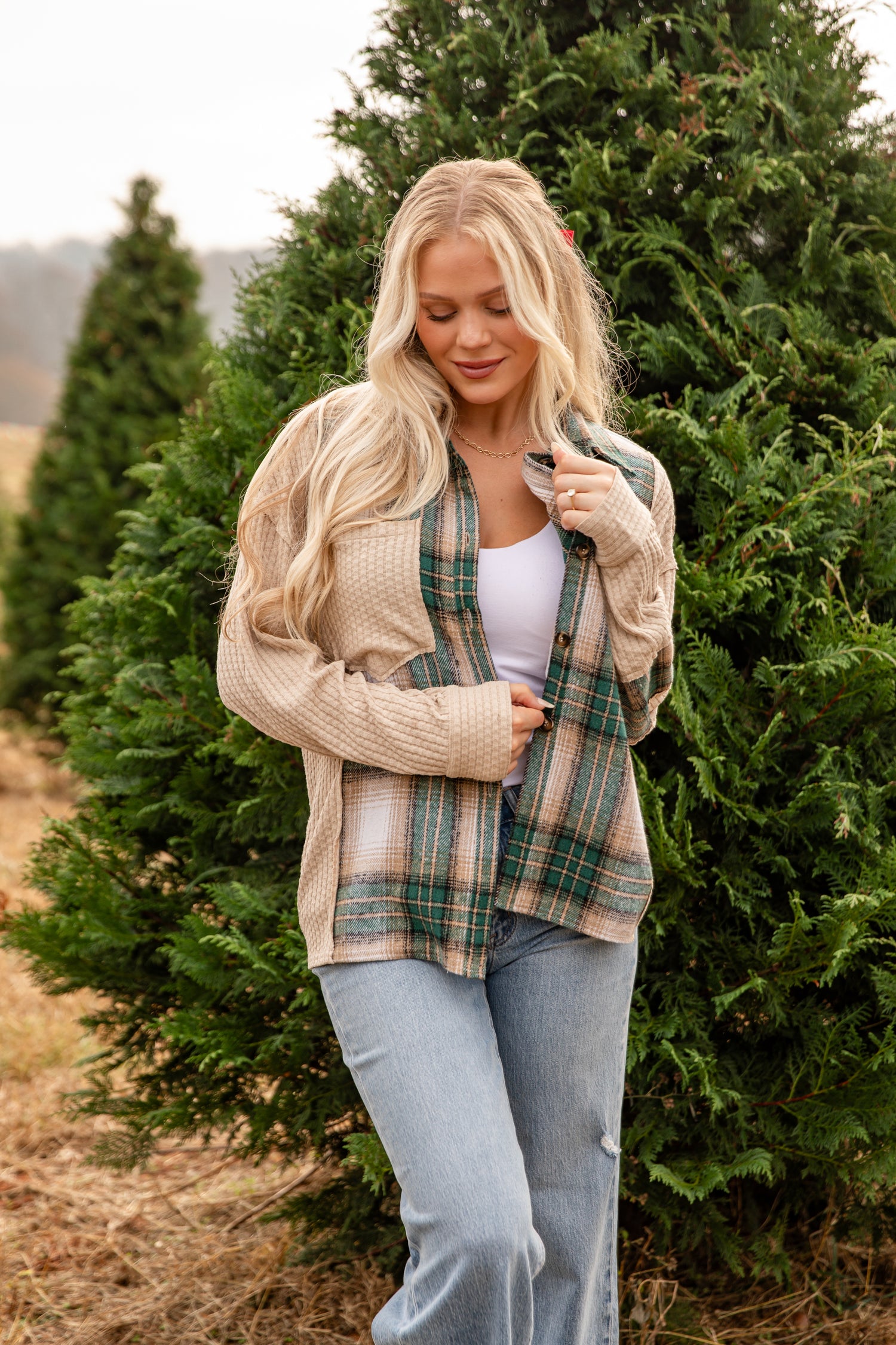 Woman standing in a field of trees wearing a plaid shirt, beige cardigan, and jeans.