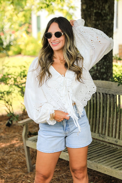 Woman wearing a white lace top and denim shorts outdoors.