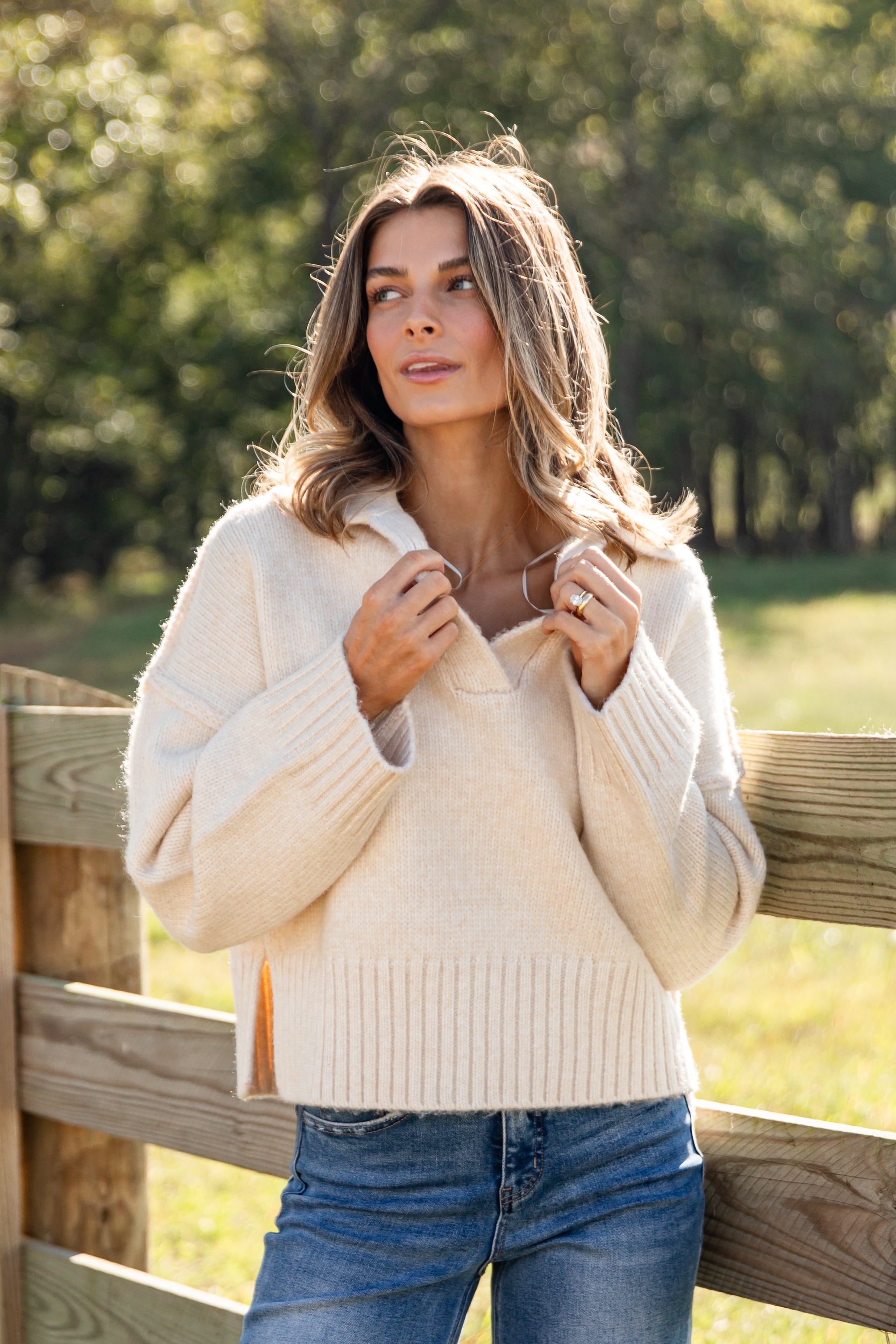 Woman wearing a cream sweater and blue jeans standing near a wooden fence with trees in the background