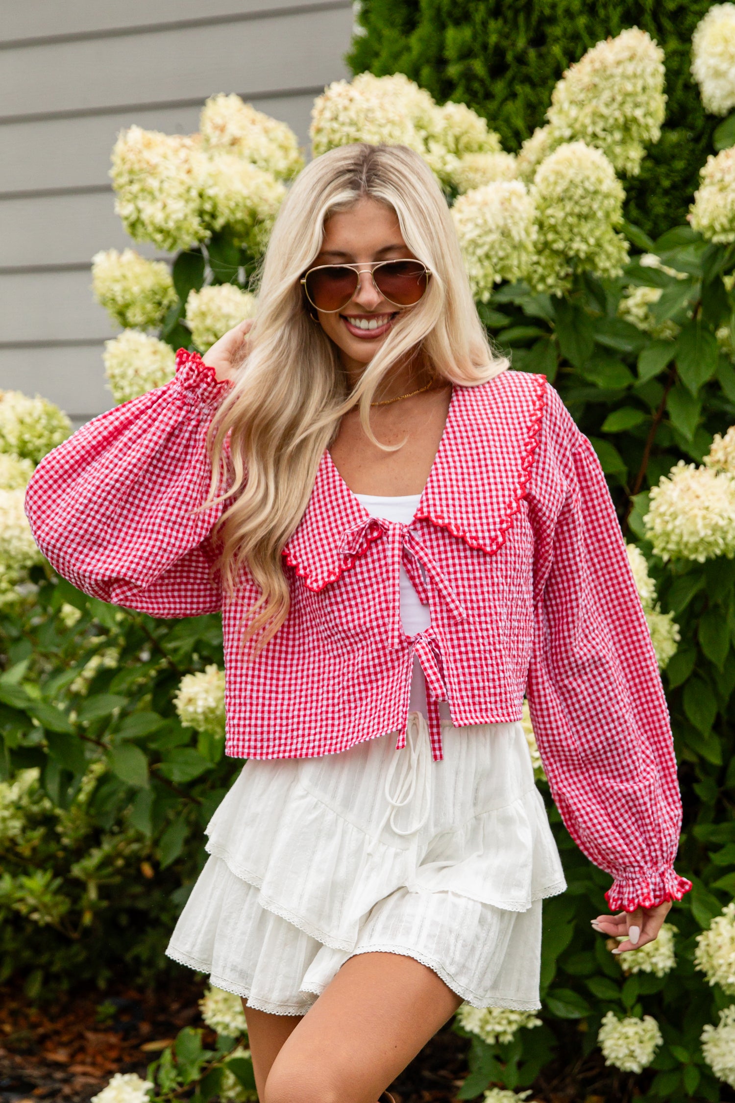 Woman wearing a red checkered blouse and white skirt standing in front of hydrangeas.