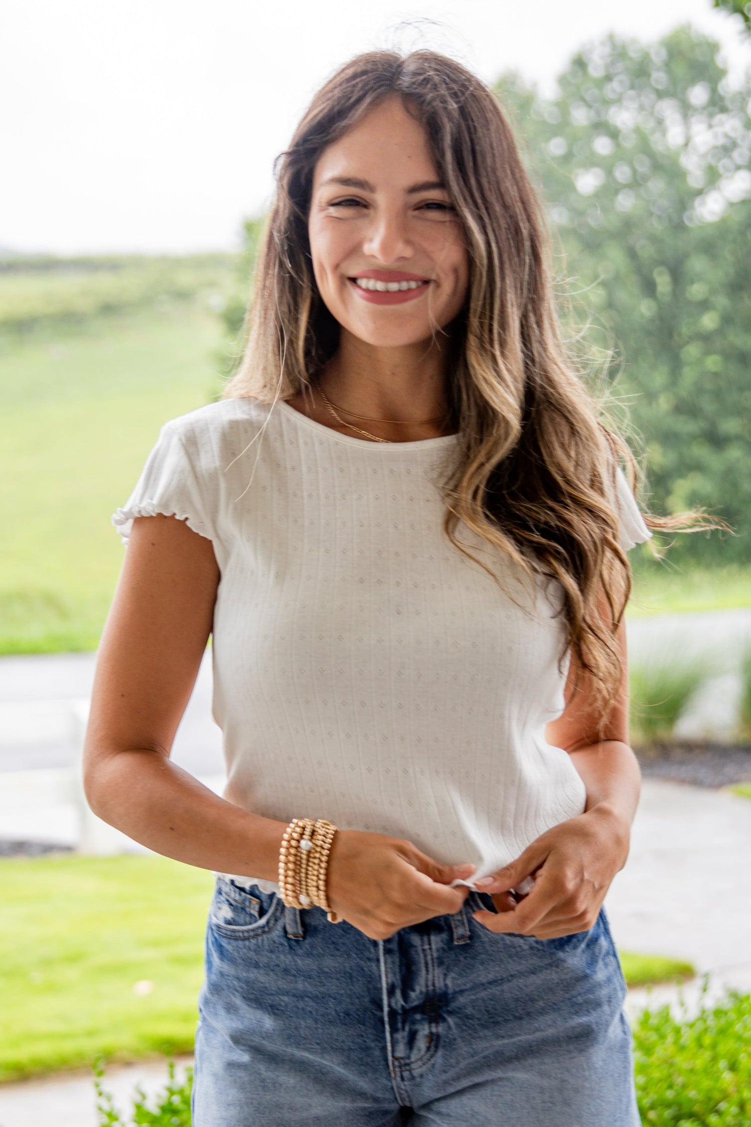 Woman standing on a porch wearing a white top, denim shorts, and beige boots with a scenic background.