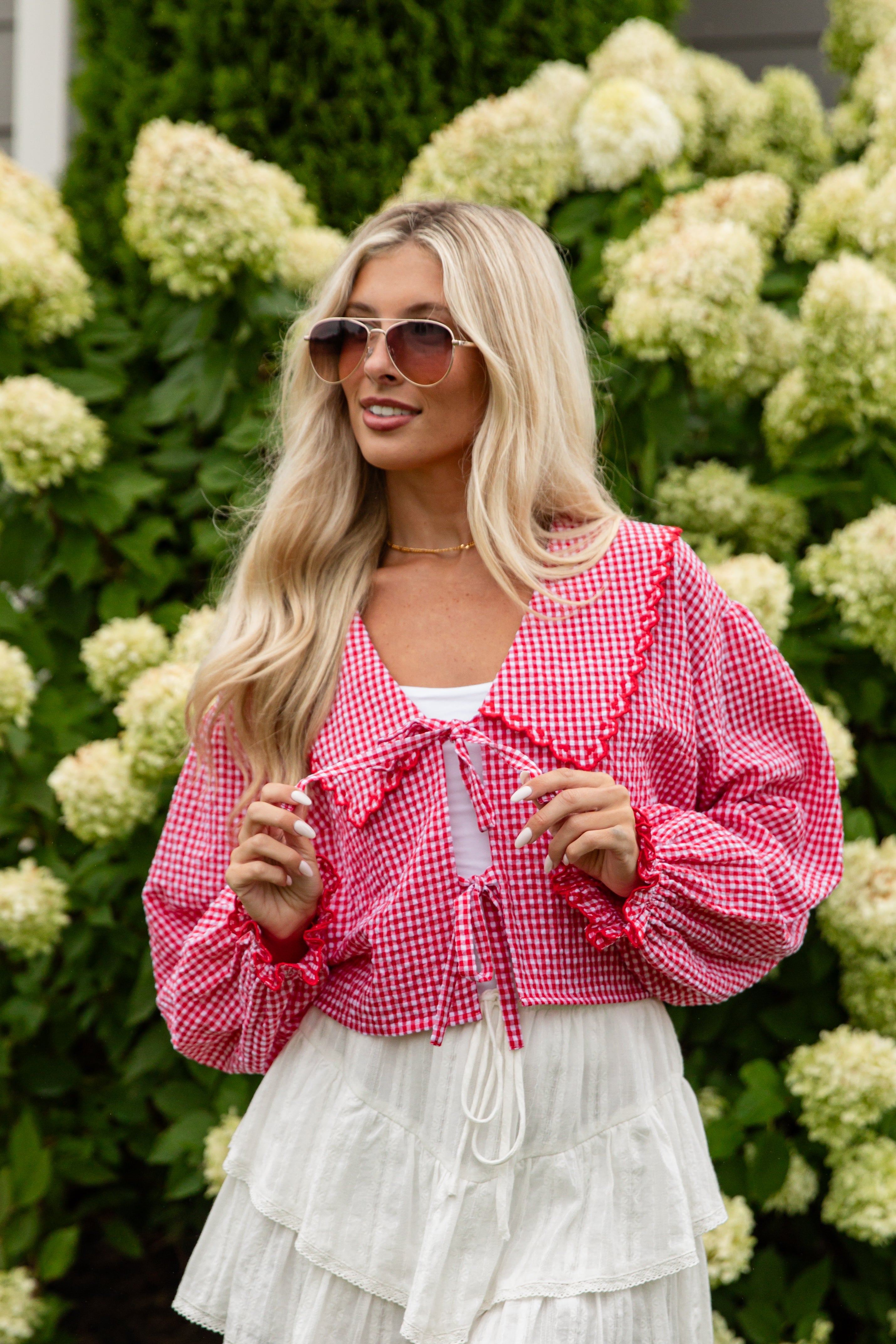 Woman wearing a pink checkered blouse and white skirt standing in front of green hydrangeas.