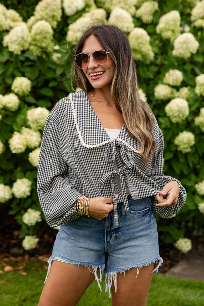 Woman wearing a checkered blouse and denim shorts standing in front of white flowers.