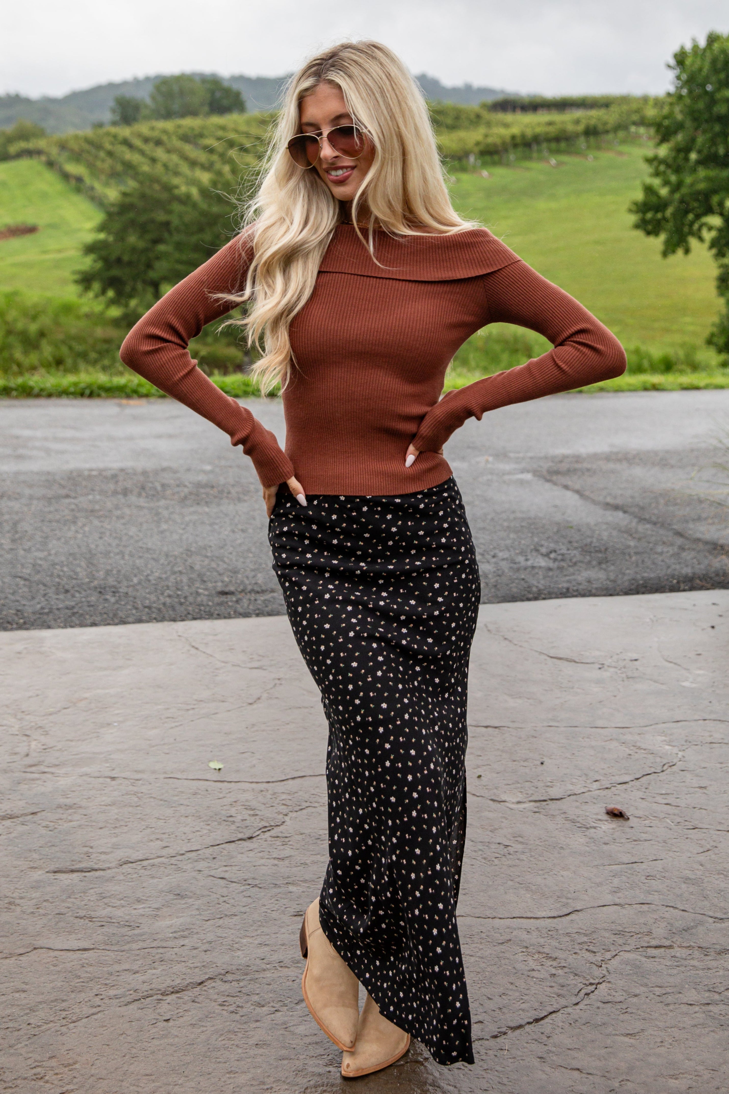 Woman wearing a brown top and black polka dot skirt standing outdoors with green fields in the background.