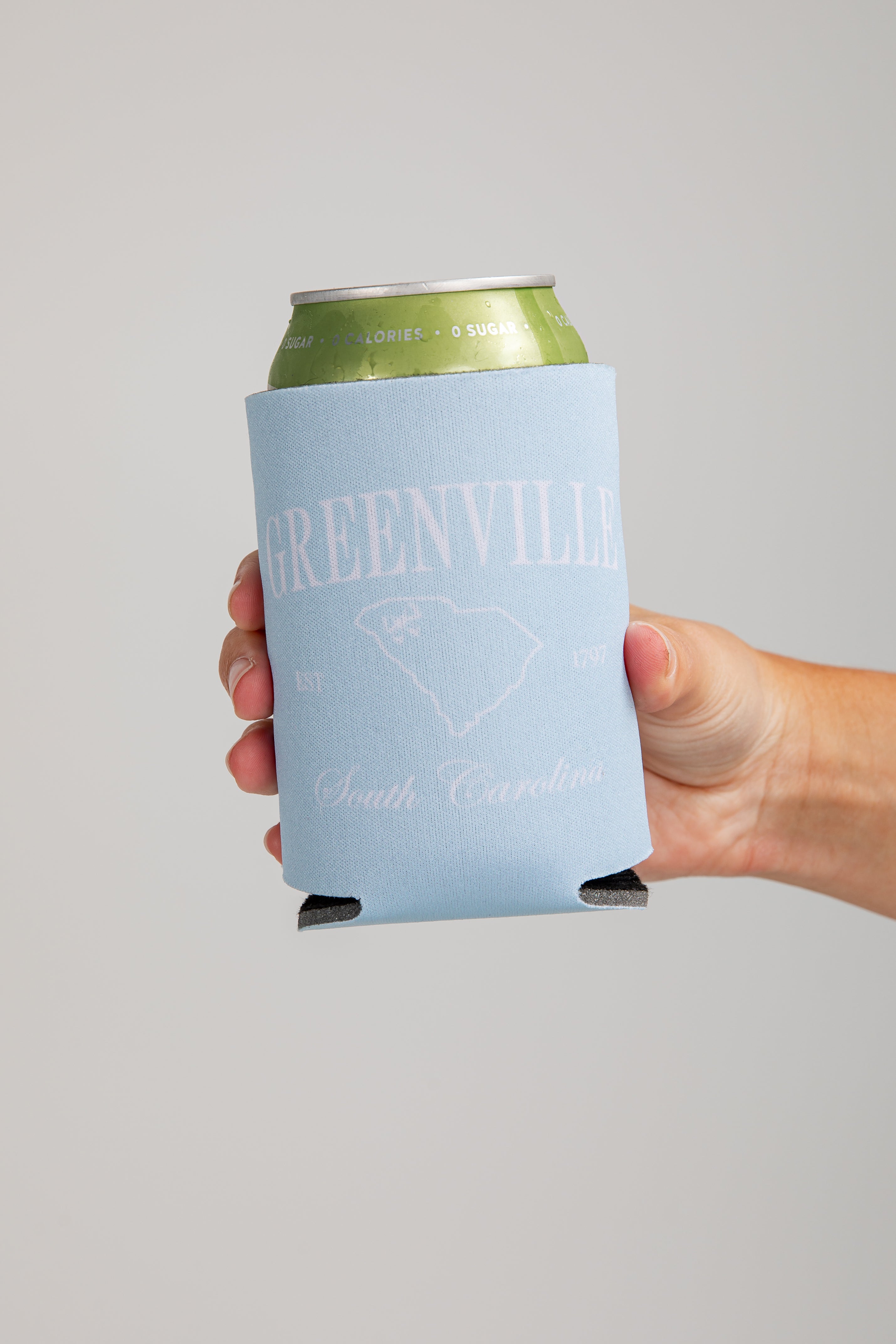 Hand holding a can with a Greenville, South Carolina branded koozie against a gray background