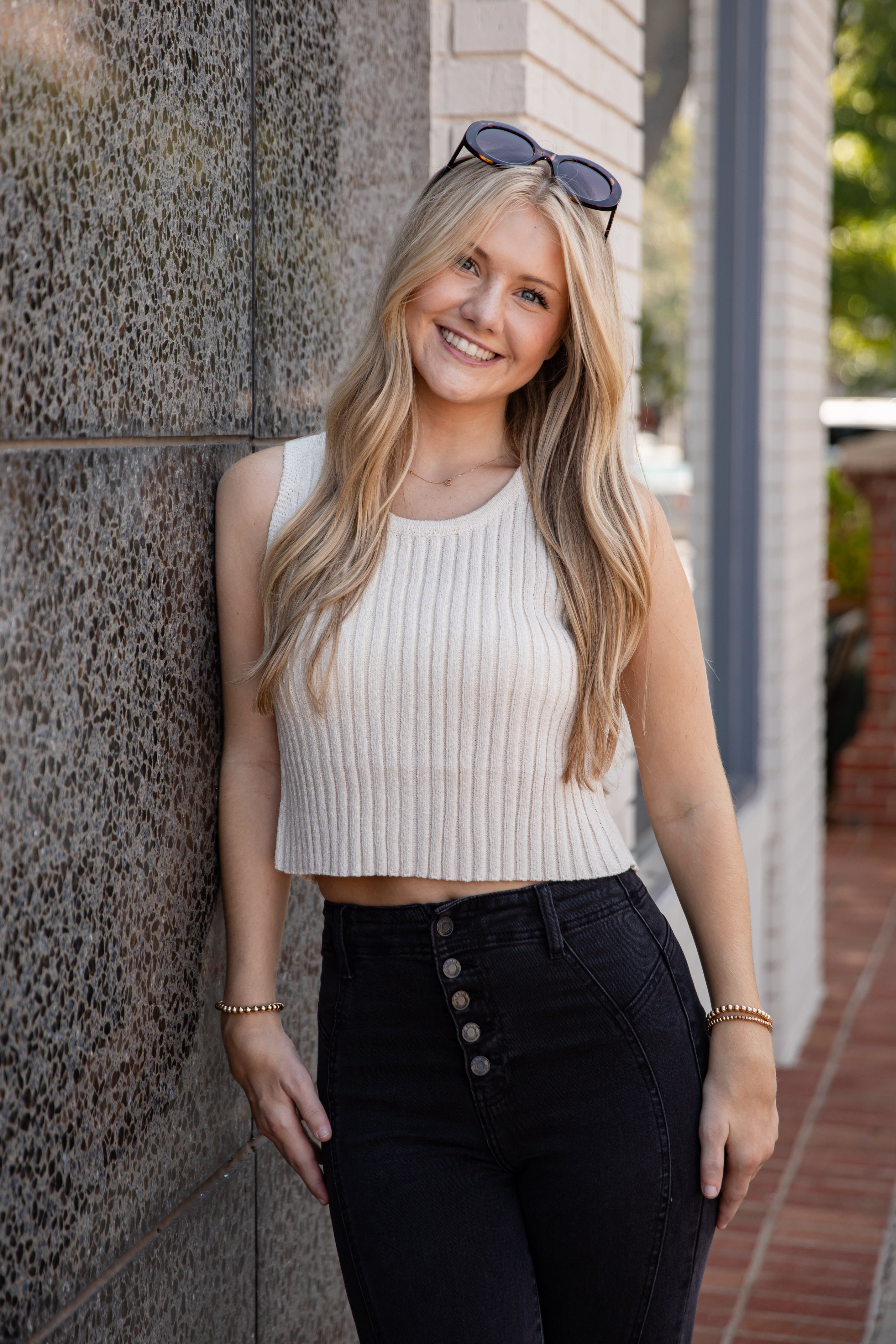 Woman in a white sleeveless top and black pants leaning against a wall.