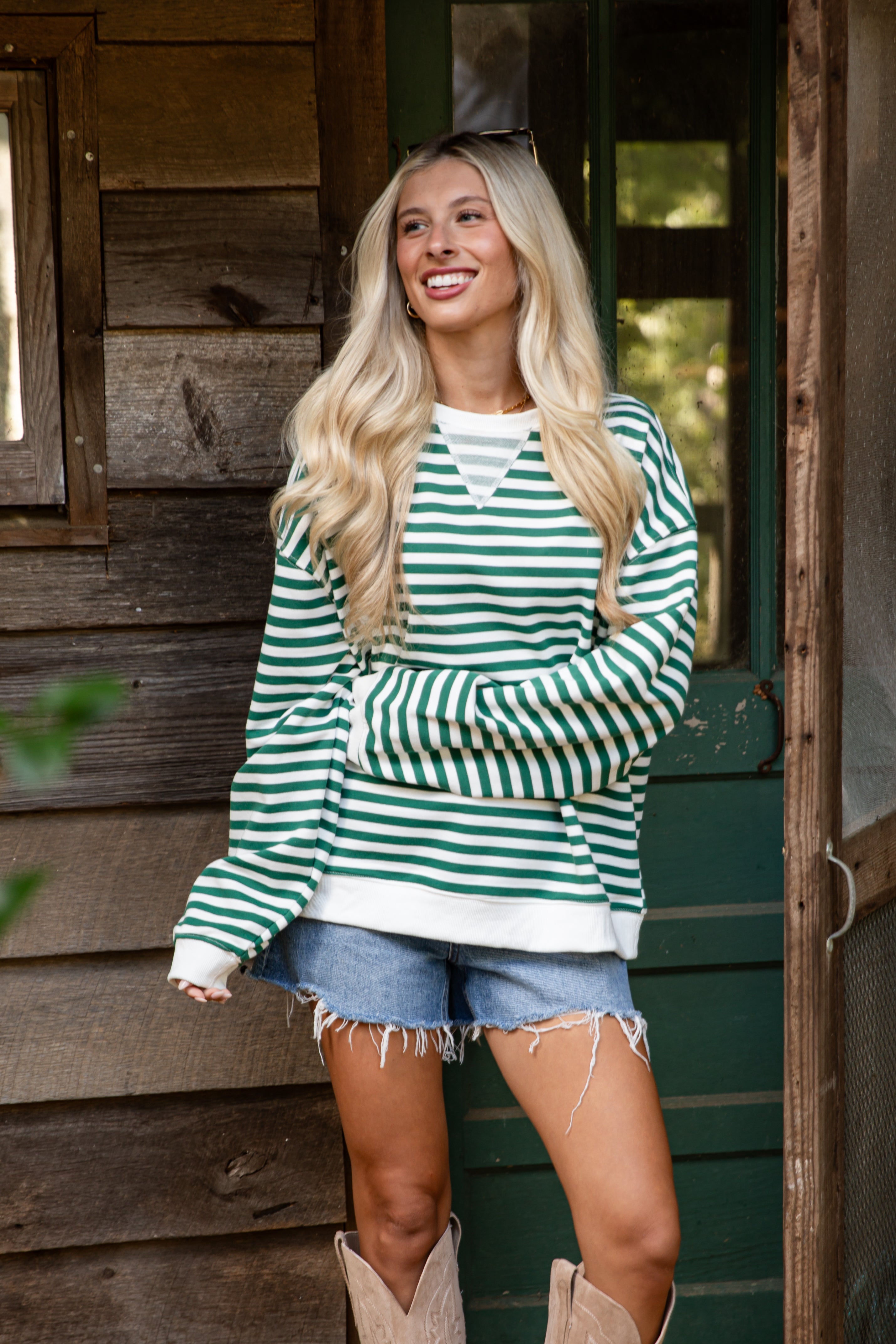 Woman wearing a green and white striped sweater and denim shorts standing in front of a wooden cabin.