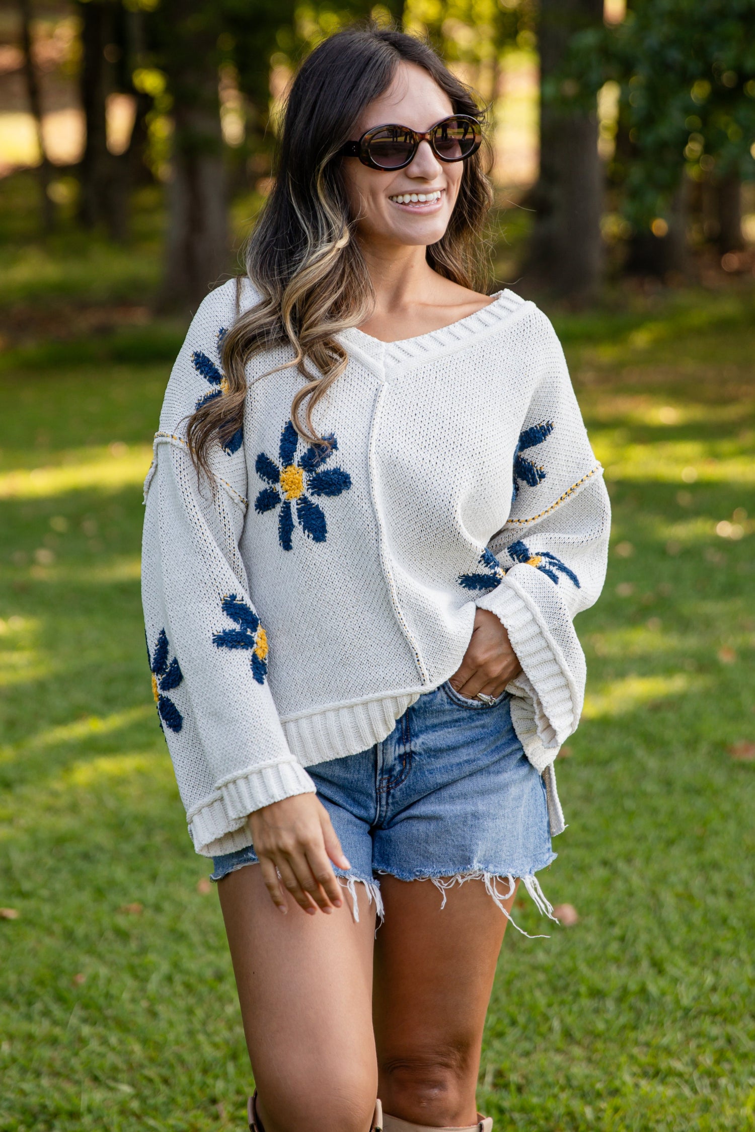 Woman wearing a white sweater with floral patterns, denim shorts, and boots in a park setting.