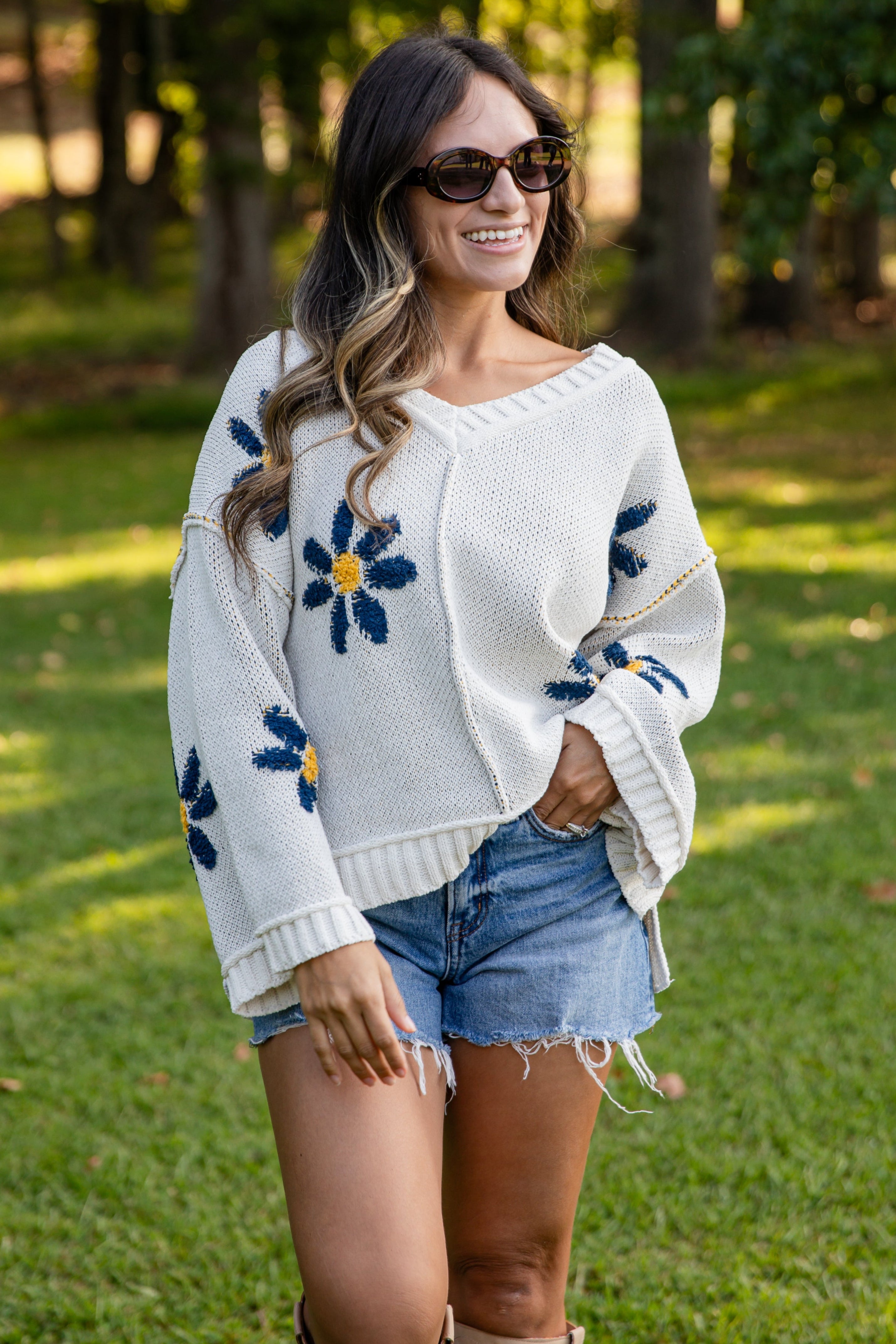 Woman wearing a white sweater with floral patterns, denim shorts, and boots in a park setting.