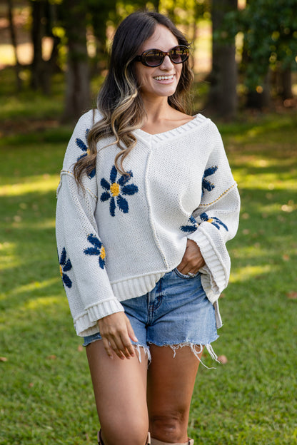 Woman wearing a white sweater with floral patterns, denim shorts, and boots in a park setting.