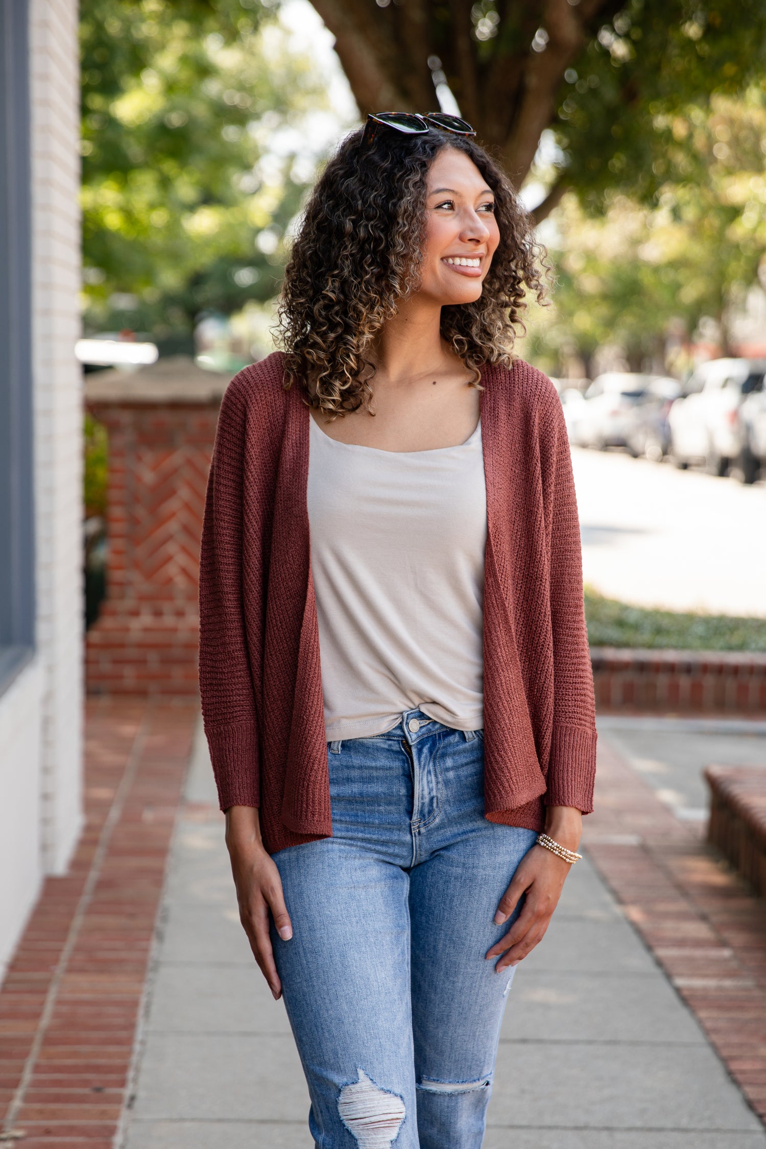 Woman wearing a rust-colored cardigan, white top, and blue jeans on a sidewalk.
