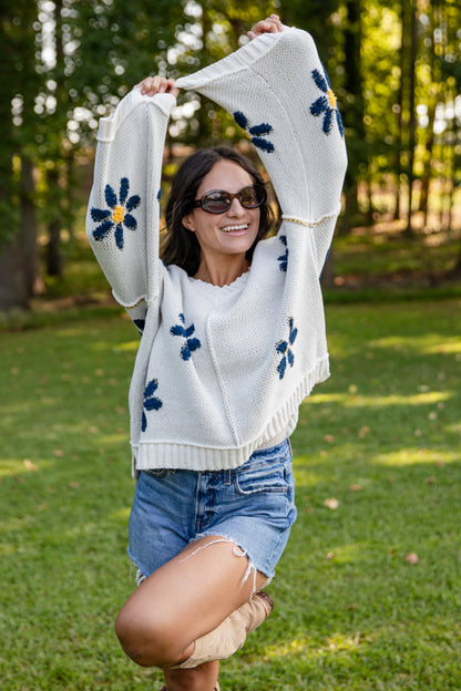 Woman wearing a white sweater with blue floral patterns in an outdoor setting