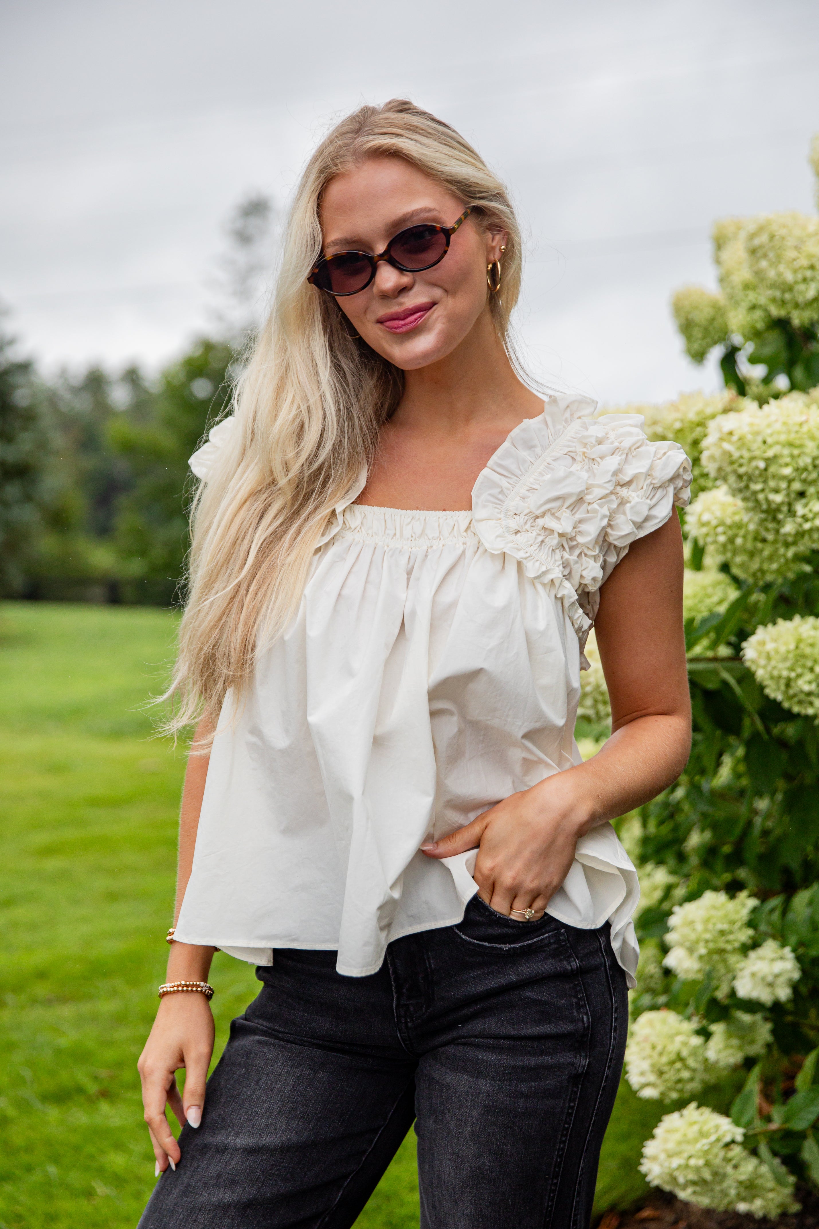 Woman wearing a white blouse with ruffled sleeves and black pants in a garden setting.