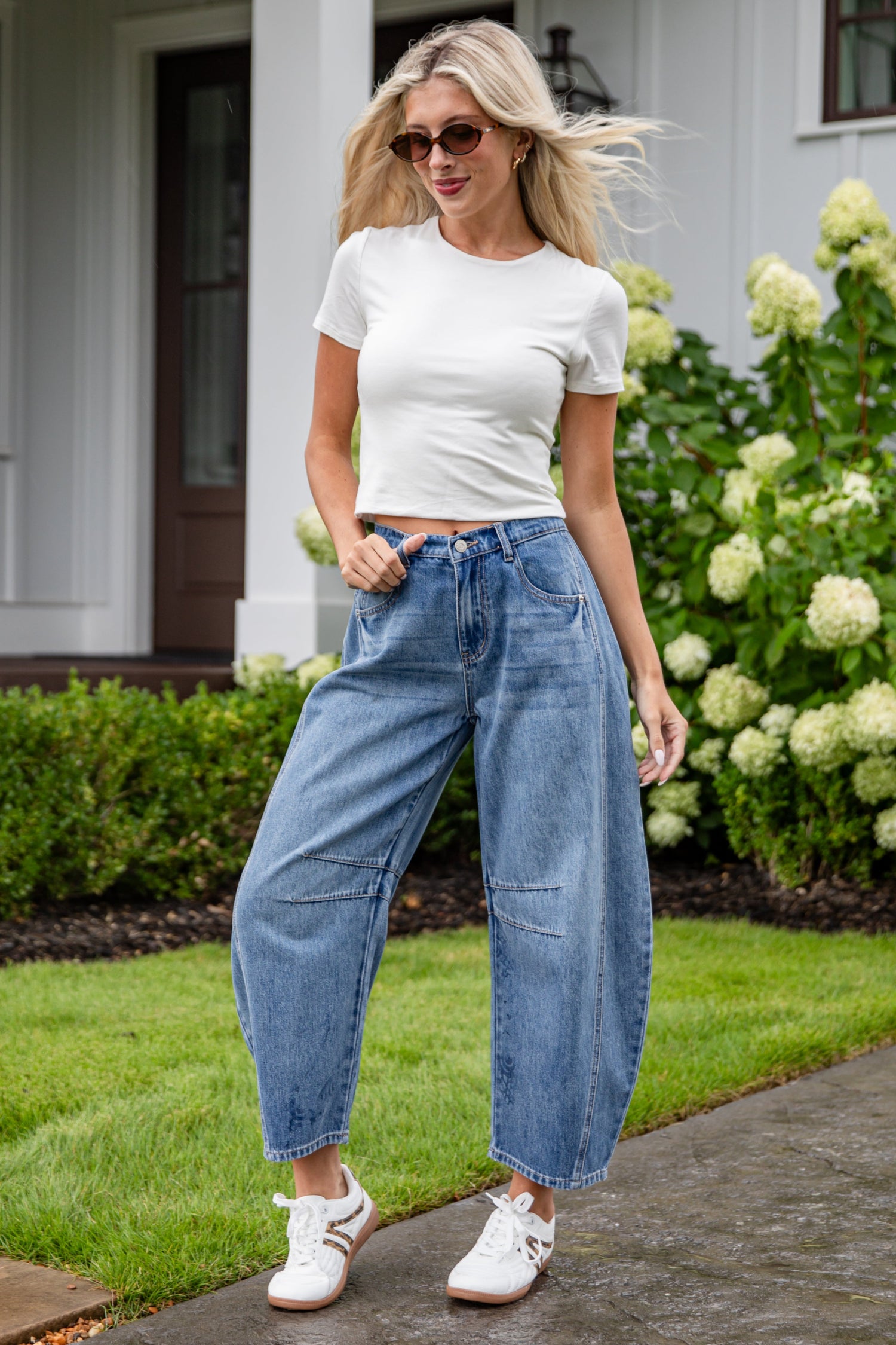 Woman wearing a white t-shirt and blue jeans standing on a sidewalk with greenery in the background