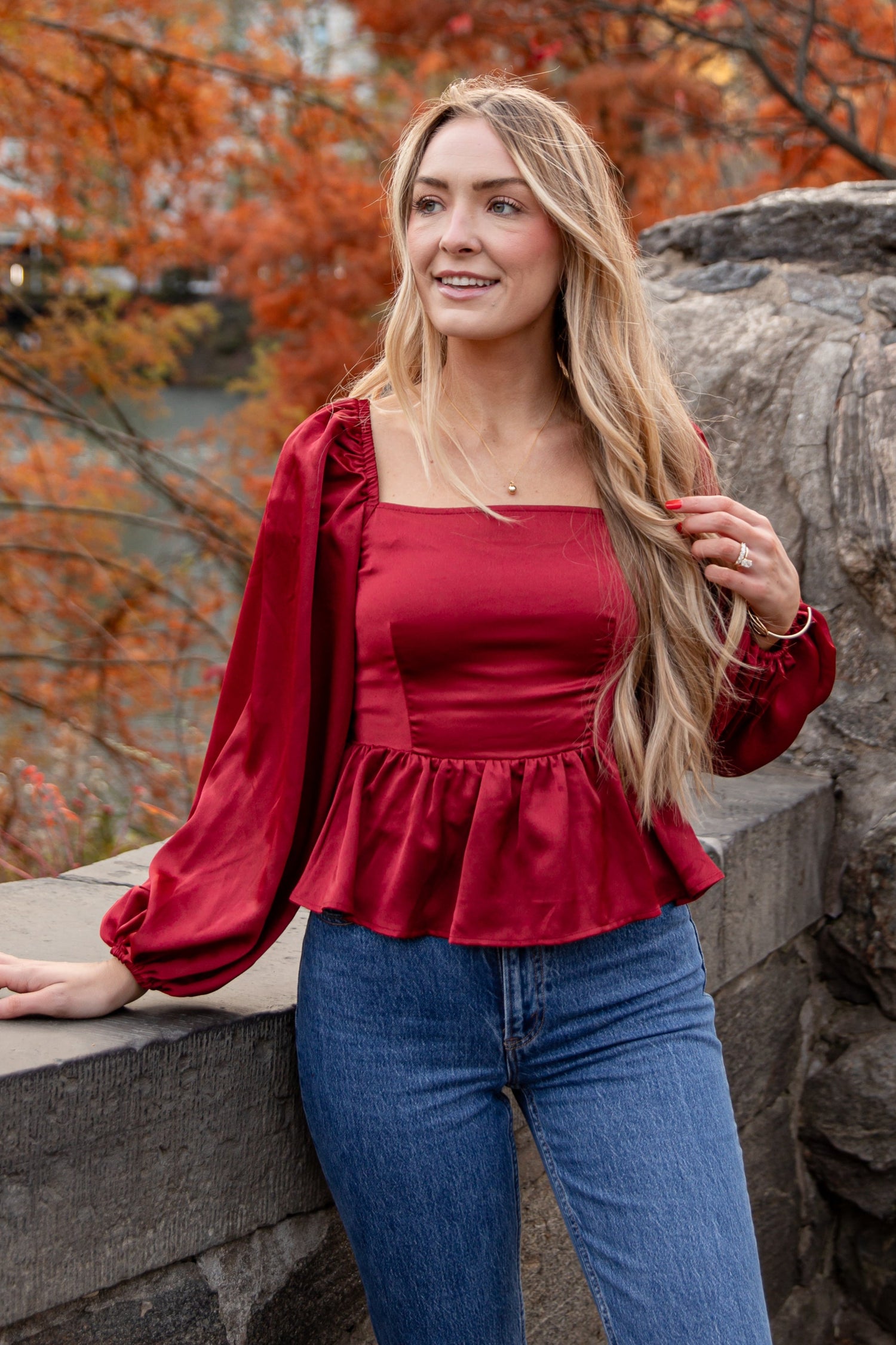 Woman in a red top and blue jeans standing in an autumn setting with trees and rocks.