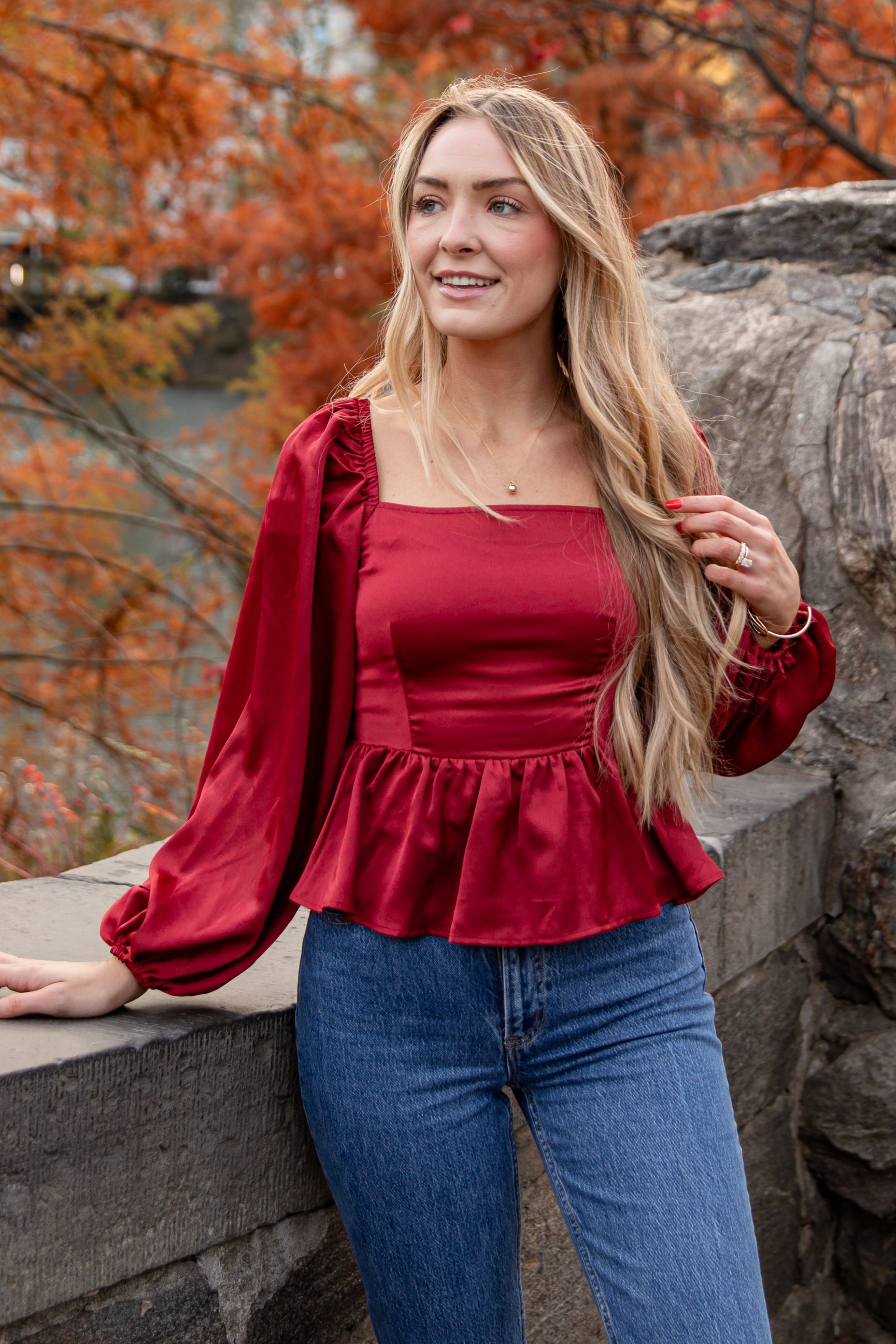 Woman in a red top and blue jeans standing in an autumn setting with trees and rocks.