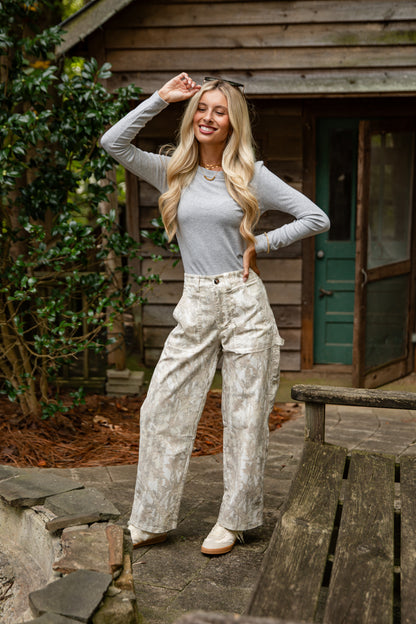 Woman standing outdoors in front of a wooden cabin, wearing a light gray top and white patterned pants.