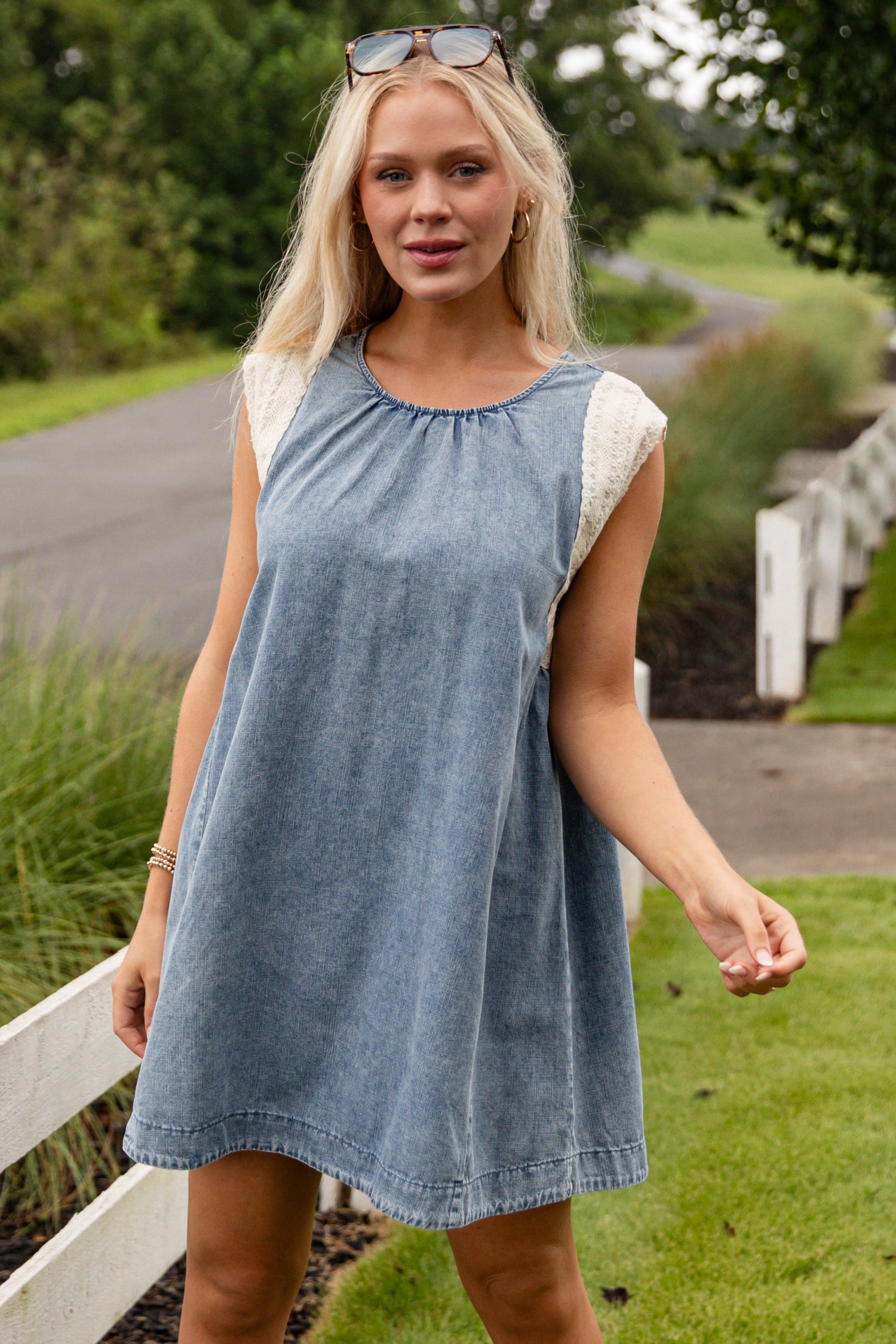 Woman in a blue dress and white boots standing on grass with a road and trees in the background
