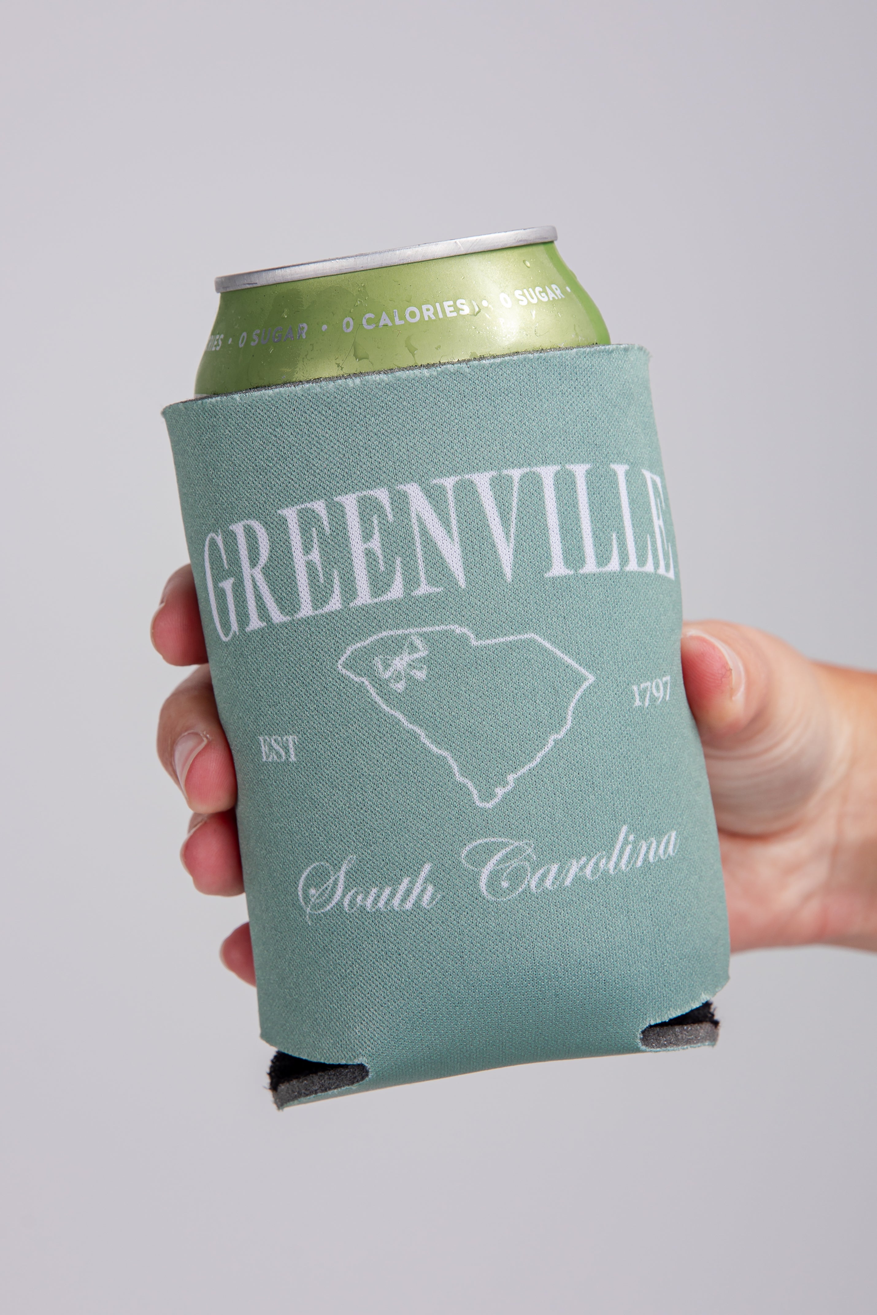 Can with a Greenville, South Carolina koozie held by a hand against a gray background