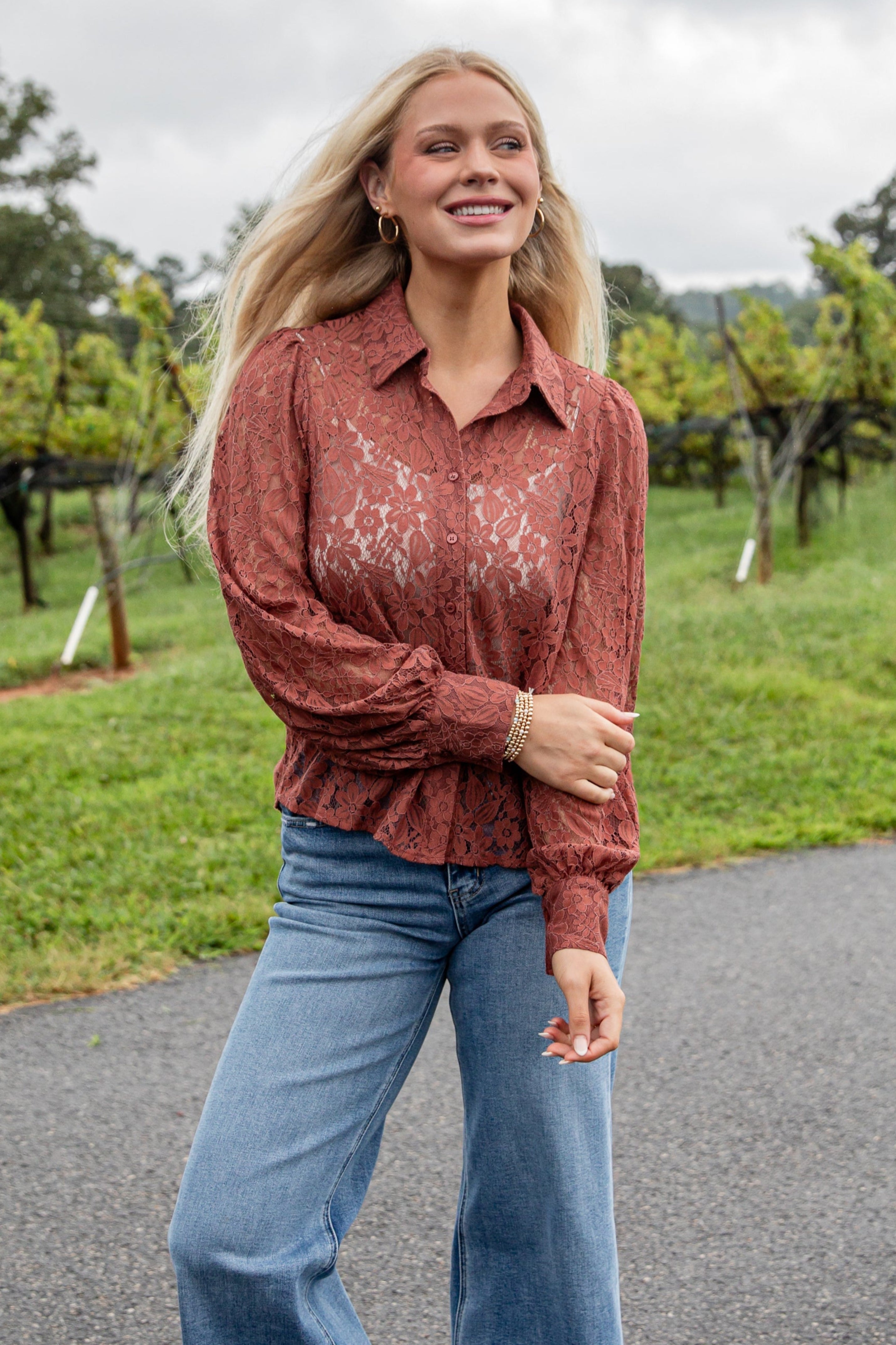 Woman in a rust-colored blouse and blue jeans standing on a road with greenery in the background